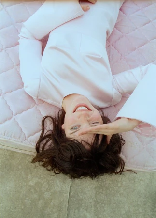 Actor Sophie Thatcher lying on a pink quilted mattress wearing a pale pink long-sleeve dress and smiling up at the camera.