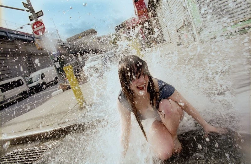 Sophie Thatcher laughing while kneeling on an asphalt city street, soaked by an explosive burst of splashing water.