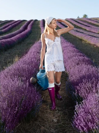 Woman in a white eyelet top and shorts walking through purple lavender rows holding a blue watering can.