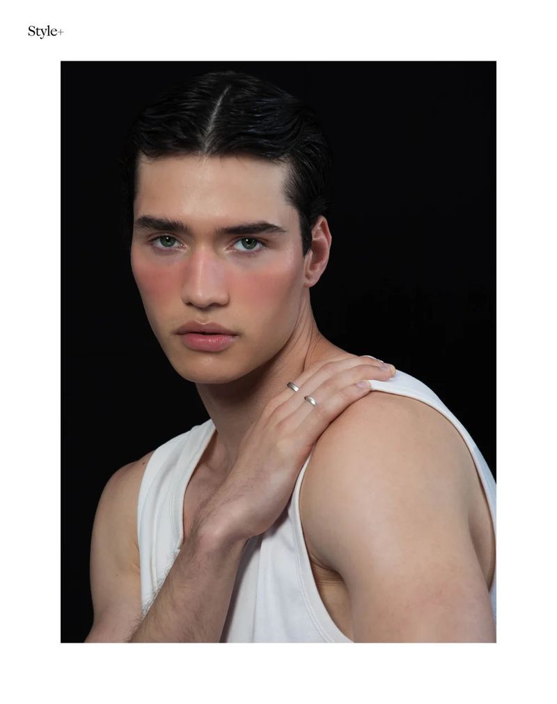 Young man with dark hair wearing a white tank top and silver rings, gazing directly at the camera against a stark black background.