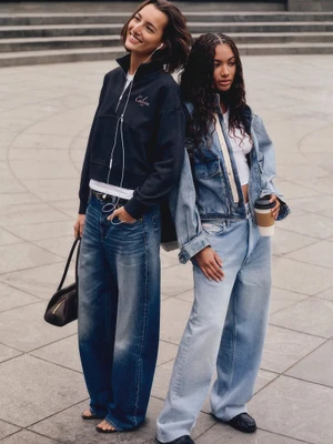 Two women posing outdoors in Calvin Klein denim and oversized casual streetwear on stone steps.
