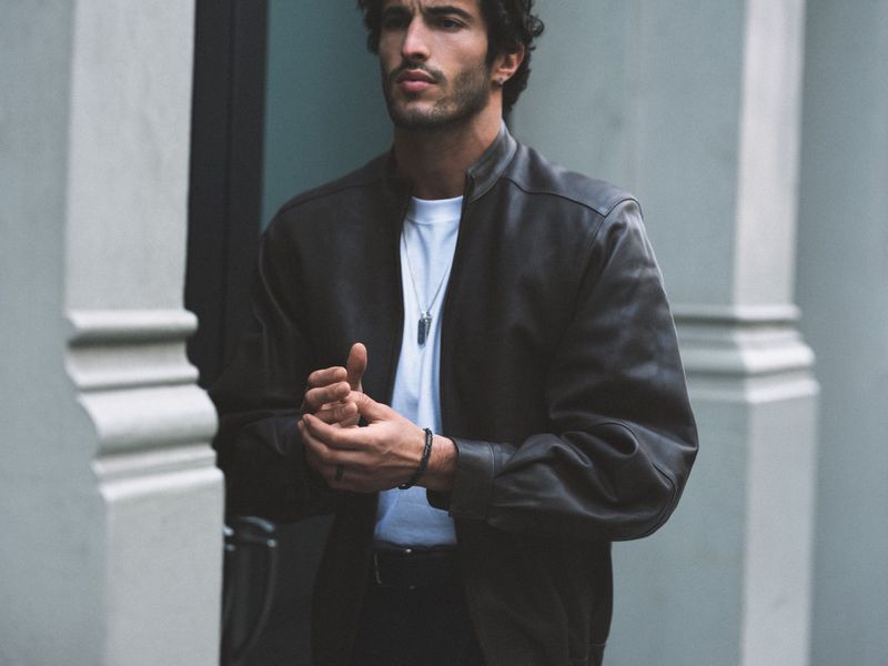 A man with dark curly hair wears a black leather bomber jacket over a white t-shirt, standing between grey architectural pillars.