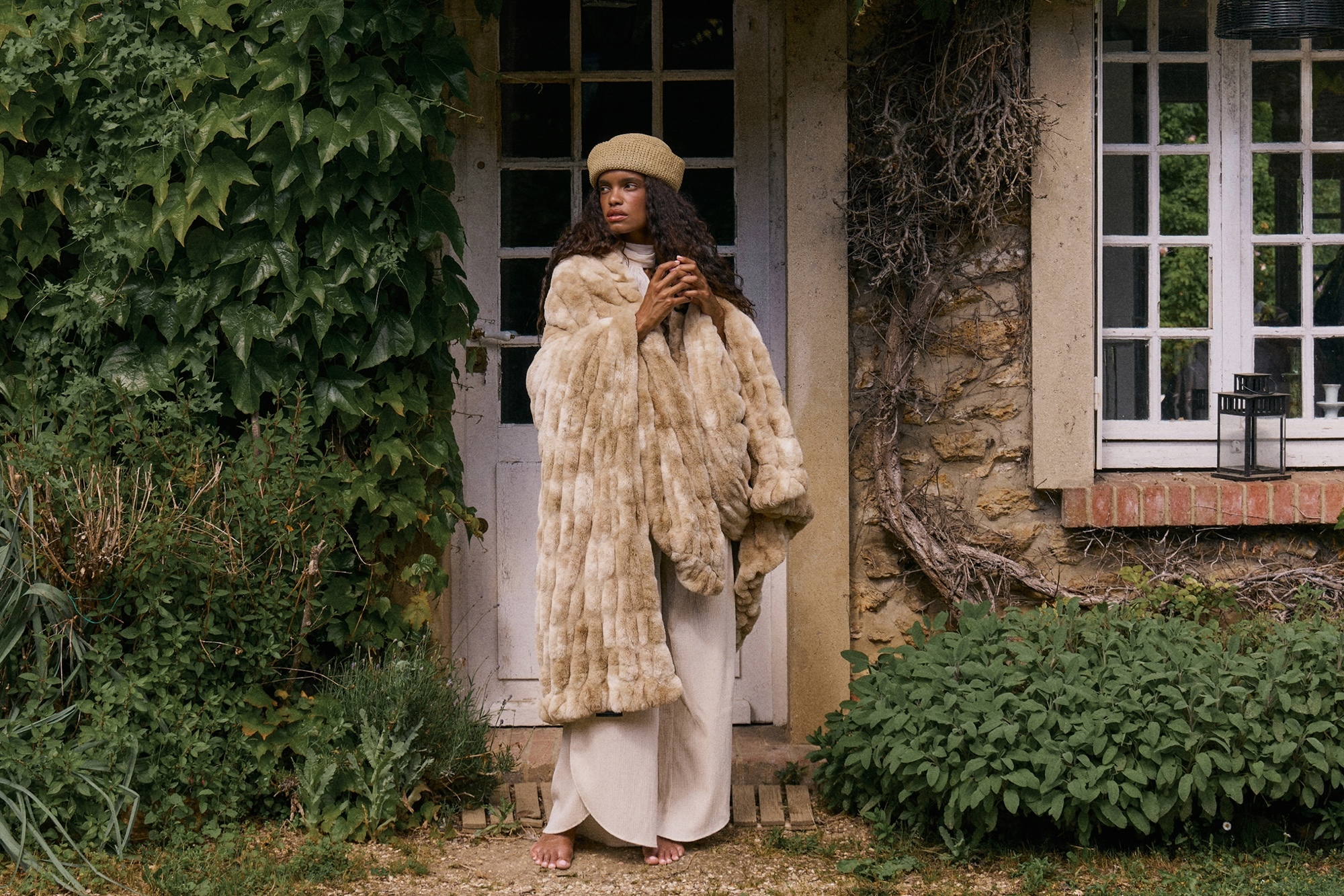 A woman with long wavy hair wearing a tan beret and textured faux-fur coat standing barefoot before a rustic stone cottage doorway.