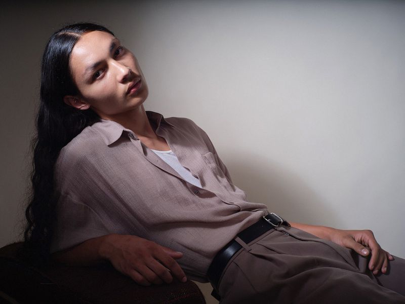 Model with long dark hair reclining in a taupe button-up shirt and pleated trousers against a neutral studio wall.