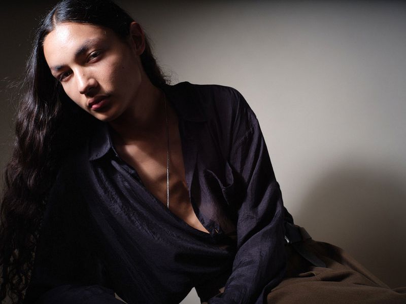 Model with long dark hair wearing an unbuttoned black sheer shirt and brown trousers against a neutral studio backdrop.