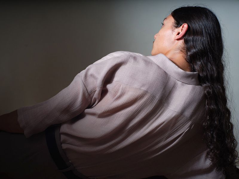 Model with long dark wavy hair seen from behind wearing a textured light taupe button-down shirt.