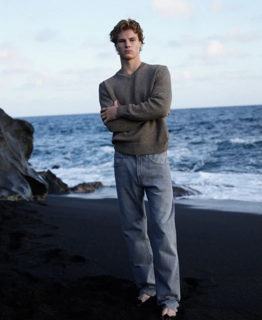 A young man with blond hair stands on a black sand beach in a khaki sweater and jeans, facing the camera, with a blue ocean backdrop.