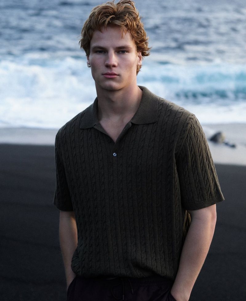 A young adult man poses in a dark green knit shirt against a backdrop of a black sand beach and ocean waves.