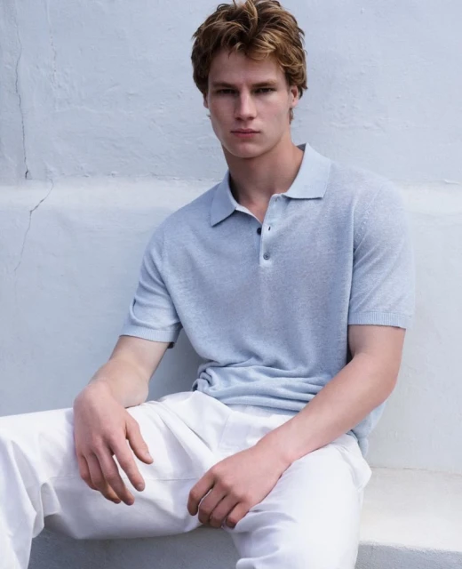 An adult male model with light brown hair poses, wearing a light blue polo shirt and white pants, seated indoors against a pale backdrop.