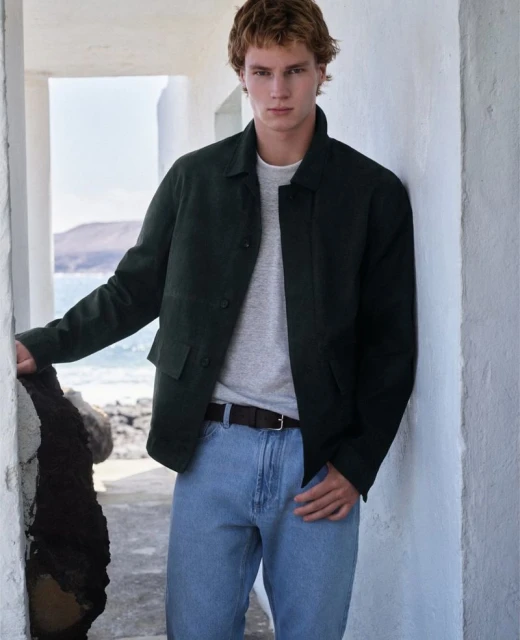 Adult male in light blue jeans and a dark green jacket posing against a white wall with an ocean background. Fashion editorial.