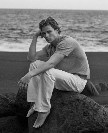 Black and white portrait of a young adult male in casual attire, seated on a rock formation with the ocean in the background.