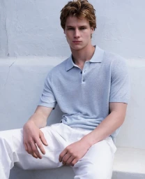 Male model seated outdoors wearing a light blue polo shirt and white trousers against a pale wall.