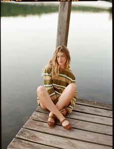 Blonde woman sitting cross-legged on a wooden dock wearing an olive striped long-sleeve top and brown strap flats by a calm lake.