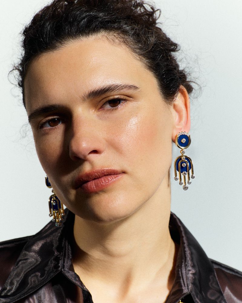 Model Iana Godnia wearing ornate gold and blue drop earrings and a dark collared shirt against a white studio background.
