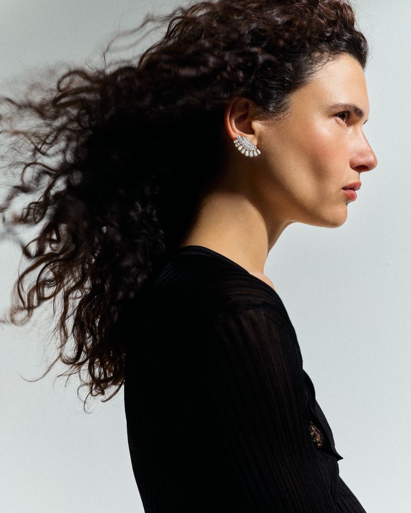 Model Iana Godnia in profile with windblown dark curls, wearing a diamond fan earring and black top against a grey seamless backdrop.