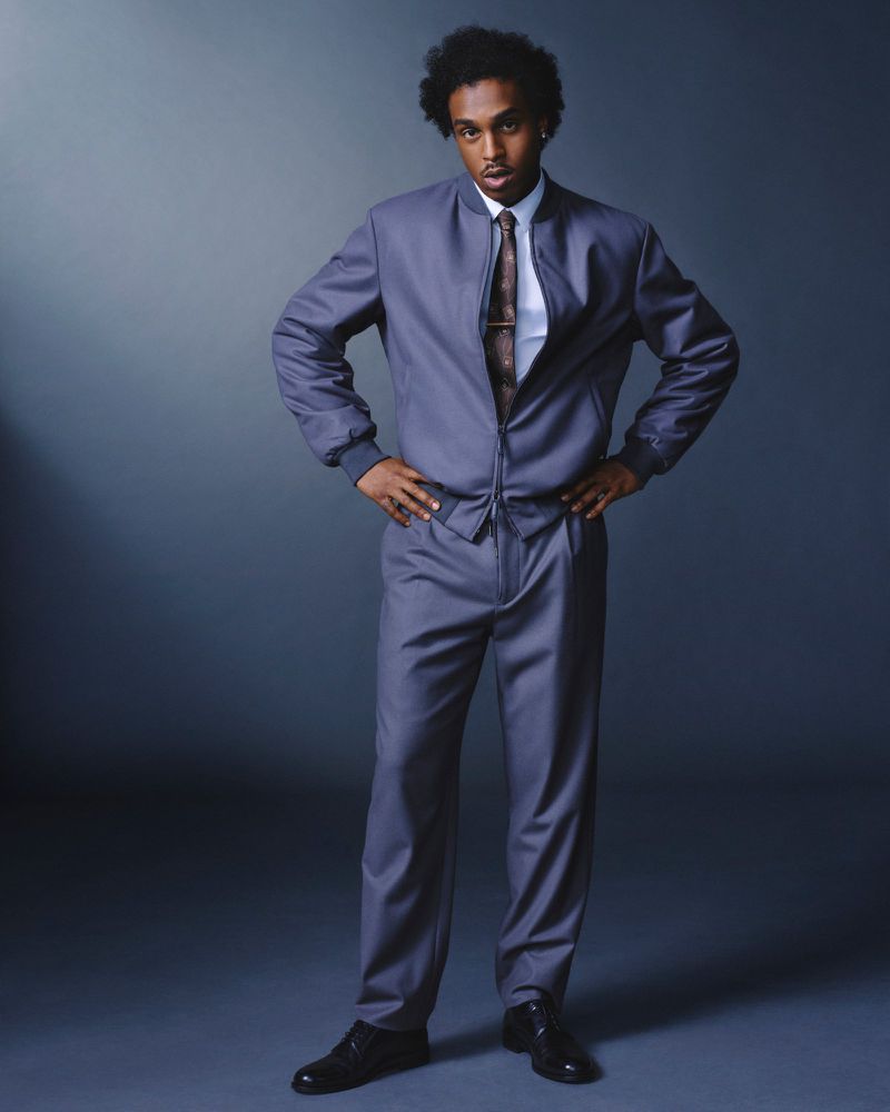 Full-length shot of an adult male with an afro, wearing a grey jacket and pants, with a tie, standing indoors in a photo studio.