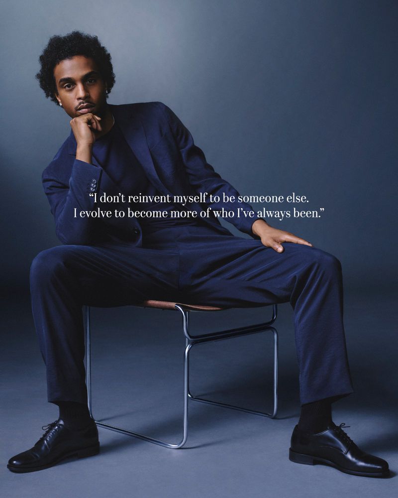Portrait of a person sitting on a chair, wearing a suit, with a hand resting on their chin, against a gray gradient background.