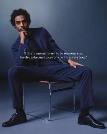 Portrait of a person sitting on a chair, wearing a suit, with a hand resting on their chin, against a gray gradient background.