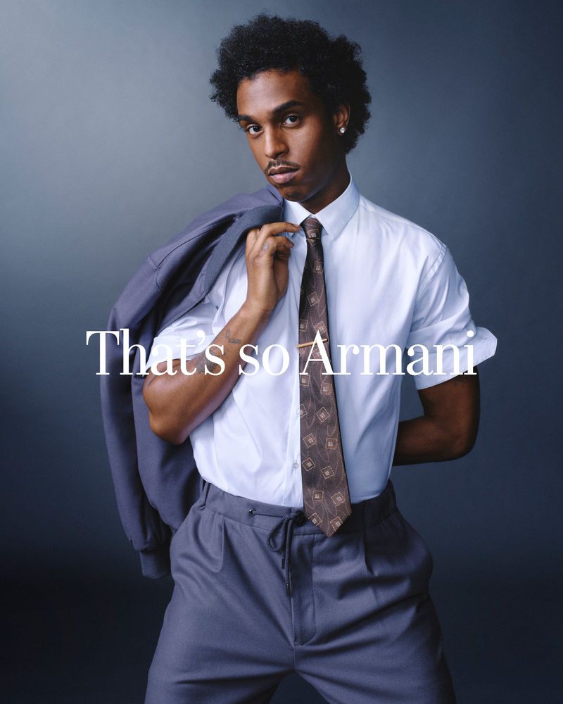 A young adult Black male in a button-down shirt, tie, and slacks poses with a jacket over his shoulder against a grey backdrop.