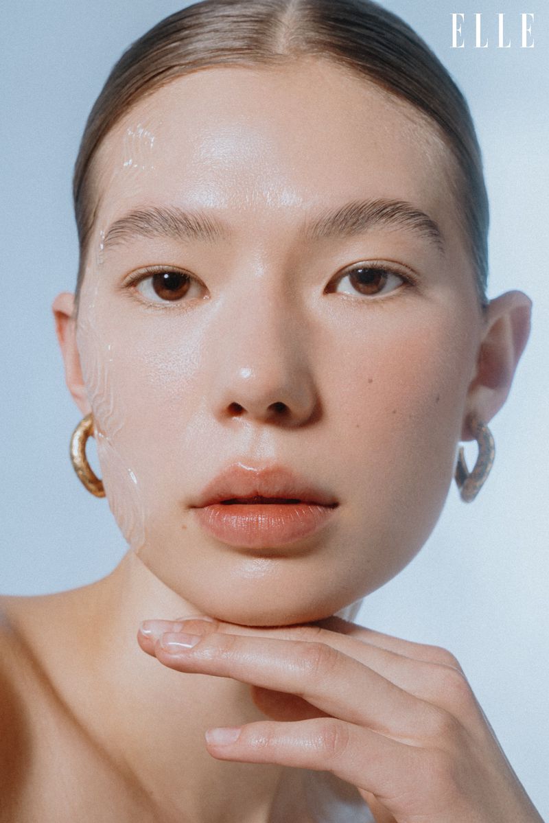 Close-up beauty portrait of an East Asian woman with dewy skin and hair tied back, posing with hand under her chin.