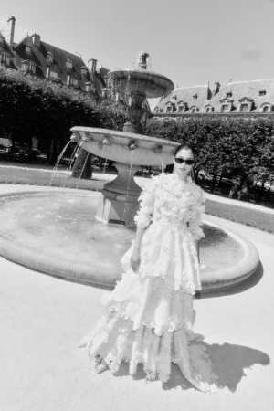 Model Mika Schneider posing in a tiered white lace couture gown and sunglasses in front of a stone fountain.