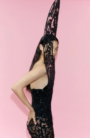 Mika Schneider wearing a black sculptural lace couture dress with a tall filigree headpiece against a pink background.