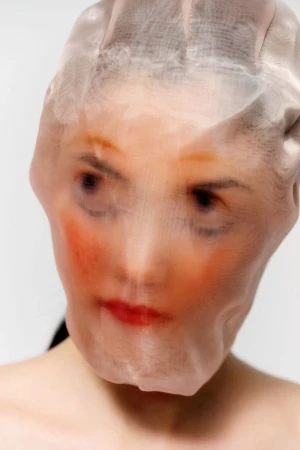 Model Mika Schneider with a sheer stocking pulled tightly over her head and face against a white studio background.