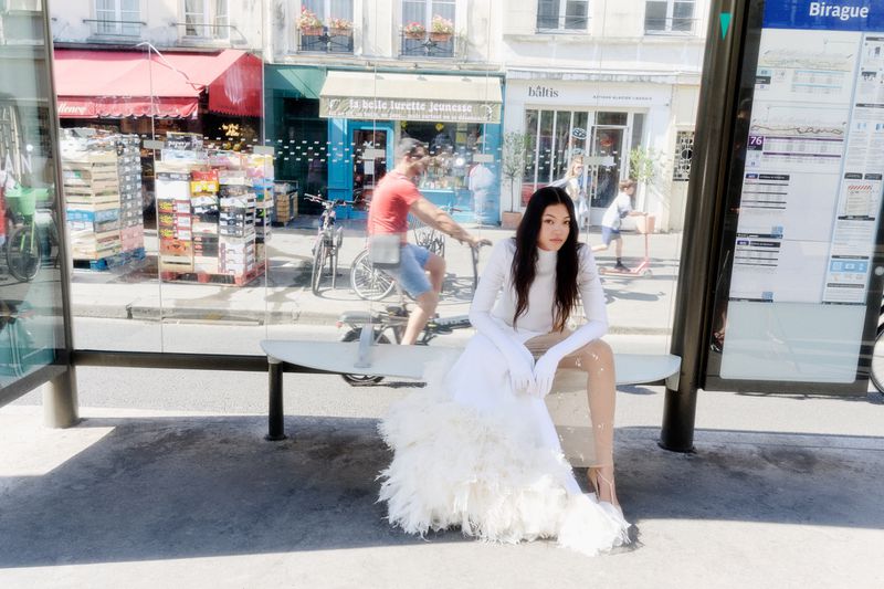 Model Mika Schneider seated at a glass bus stop bench wearing a white long-sleeved feather gown in Paris.