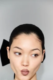 Close-up portrait of model Mika Schneider making a playful pout face with a black hair bow against a grey backdrop.