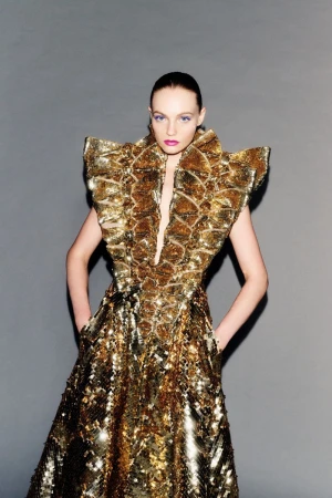 Fran Summers wearing a sculptural metallic gold sequin dress with exaggerated shoulders against a plain grey studio backdrop.