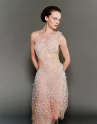 Model Fran Summers wearing a sheer pearl-embellished one-shoulder dress against a neutral grey studio backdrop.