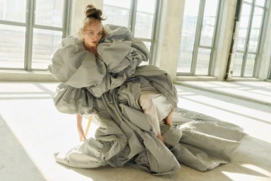 Model Fran Summers seated in an oversized voluminous grey sculpted gown in a sunlit industrial loft space.