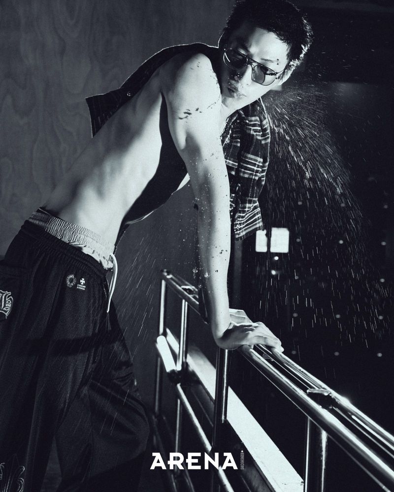 Shirtless East Asian man in sunglasses and black track pants leaning on a metal railing amid flying water droplets.