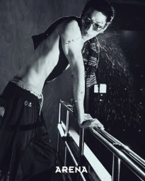 Shirtless East Asian man in sunglasses and black track pants leaning on a metal railing amid flying water droplets.