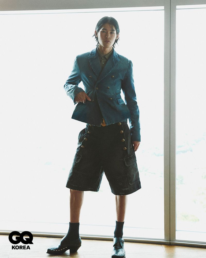 Model Han Jin Hee wearing a double-breasted denim blazer and dark shorts standing before large windows for GQ Korea