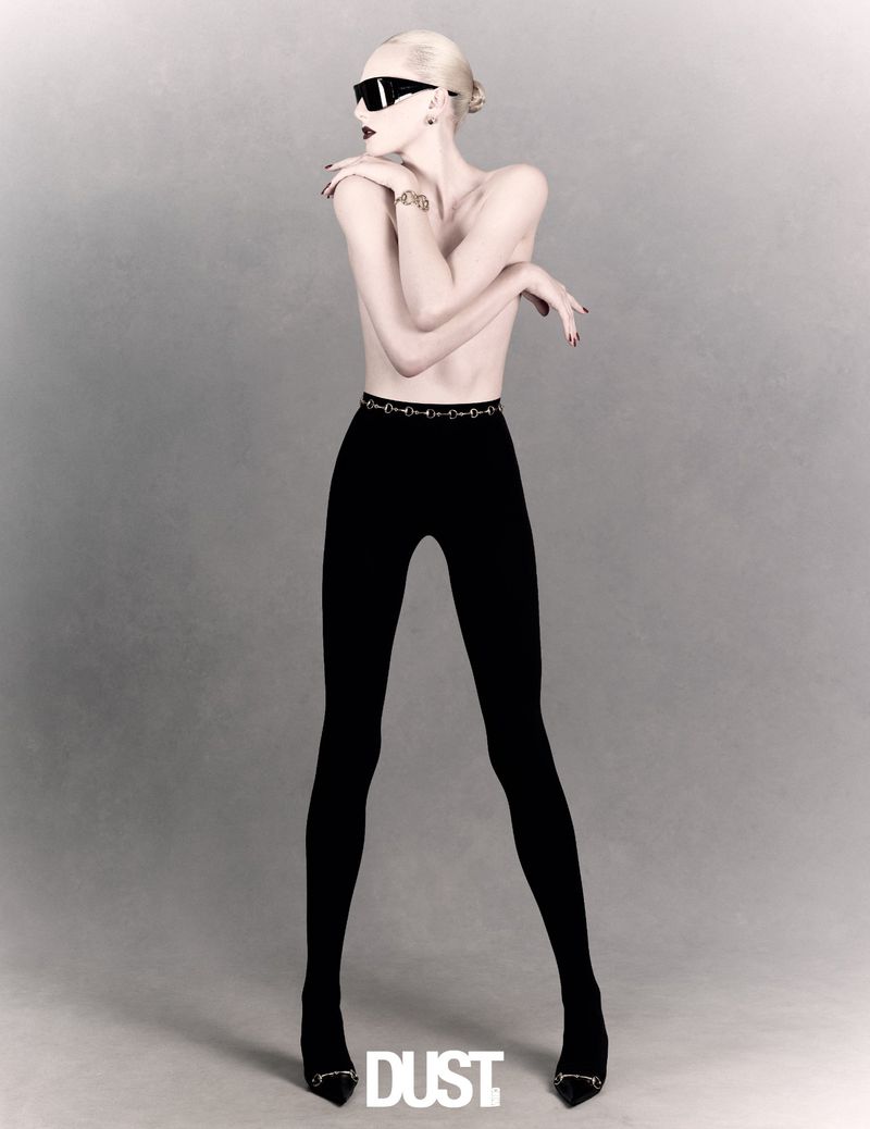 Alex Consani wears black leggings, pointed heels, and wraparound sunglasses against a soft, neutral grey studio background.