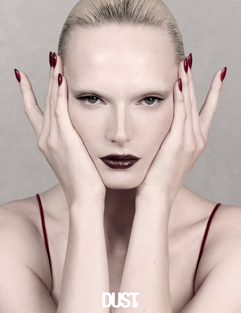 Alex Consani with bleached eyebrows and dark red lipstick, holding hands to face with matching dark red manicure against grey background.