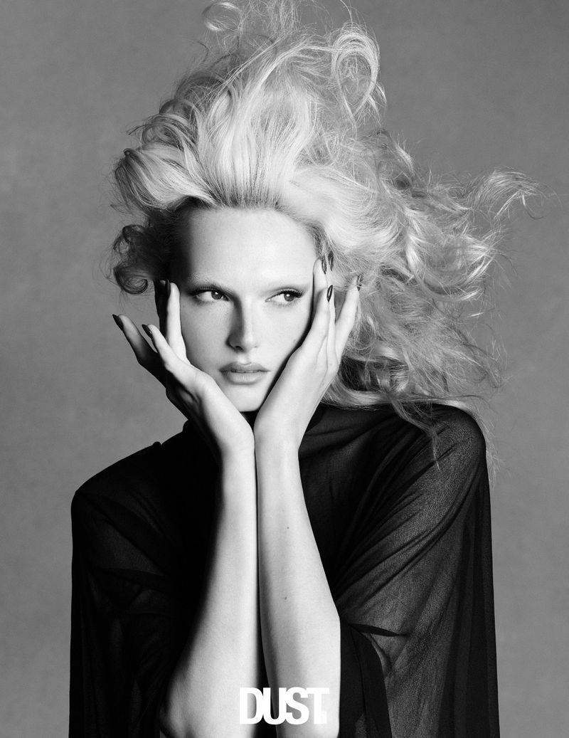 Alex Consani with voluminous blonde hair and bleached eyebrows posing with hands on face in a black top against a grey background.