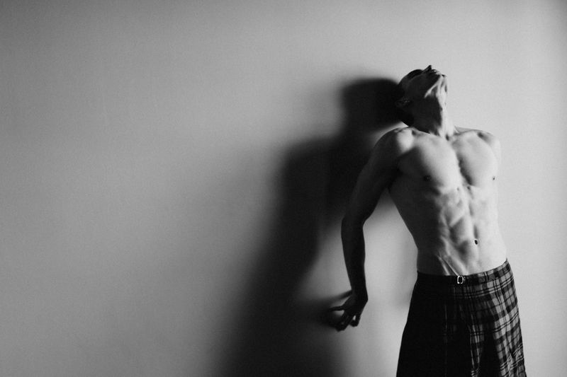 Shirtless male model in a plaid kilt leaning back against a wall in high-contrast black and white.