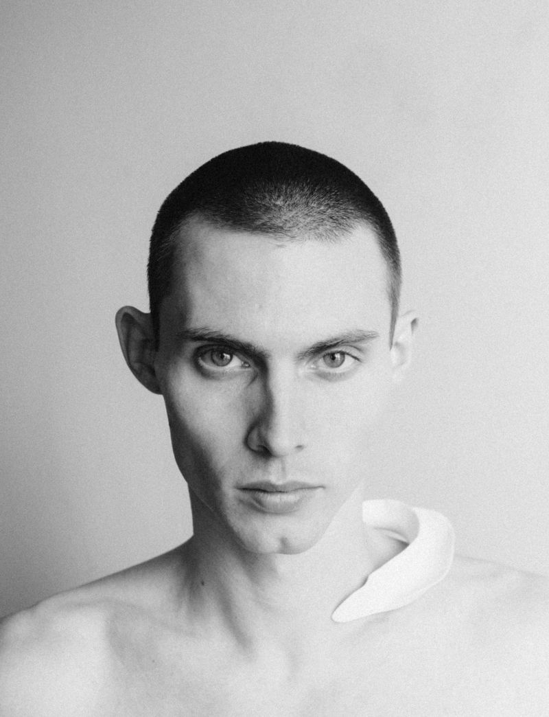 Black and white portrait of model Arnau Sarda with a buzz cut and a curved white sculptural collar piece against a plain background.