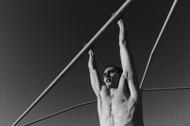 Shirtless model Arnau Sarda hangs by his hands from an outdoor metal bar against an open sky in black and white.