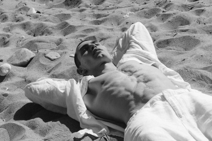 Arnau Sarda lying on a sandy beach wearing an unbuttoned white shirt and trousers in a black and white editorial.