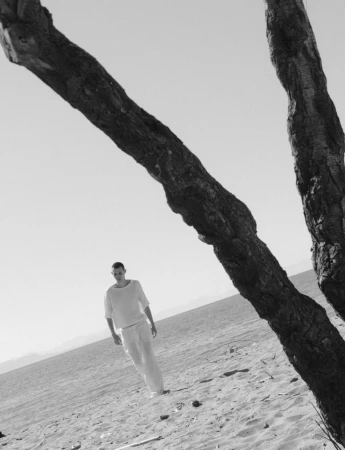 Arnau Sarda walks across a sandy beach in white clothing, framed by a large weathered tree branch in black and white.