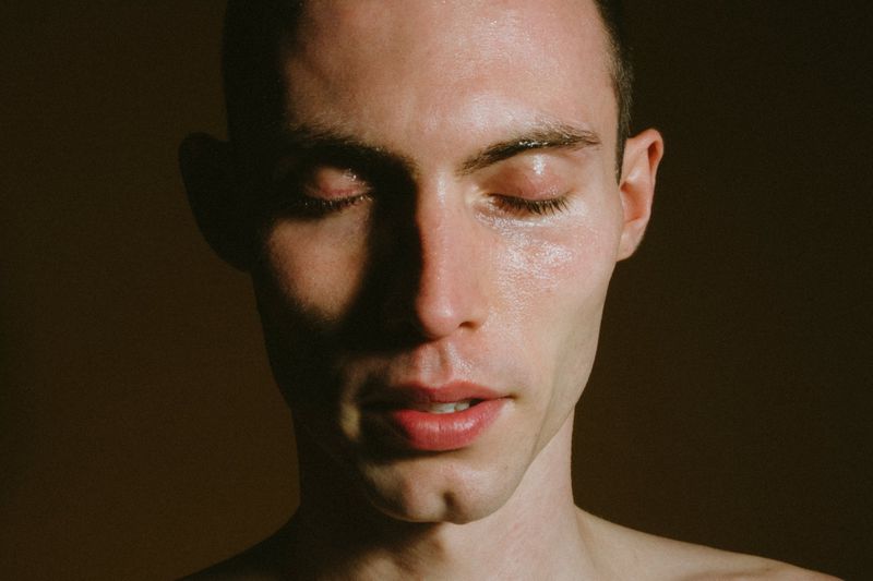 Close-up portrait of model Arnau Sarda with eyes closed and dewy skin against a dark muted backdrop.