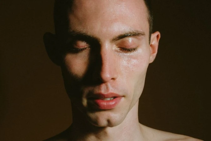 Close-up portrait of model Arnau Sarda with eyes closed and dewy skin against a dark muted backdrop.