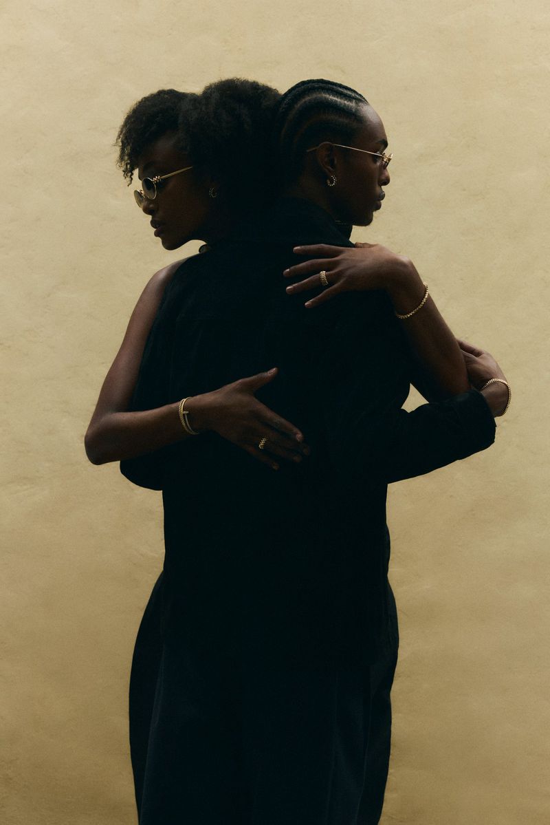Two Black models in black attire standing back to back in an intimate embrace against a warm beige textured backdrop.