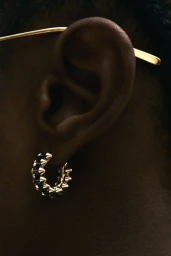 Close-up macro view of a Black model's ear wearing a gold studded hoop earring and thin gold eyewear temple.