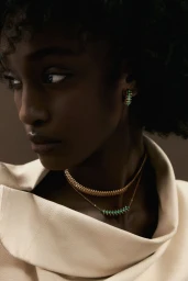 Close-up of a Black woman model wearing layered gold and green gemstone necklaces with a matching earring and cream draped top.