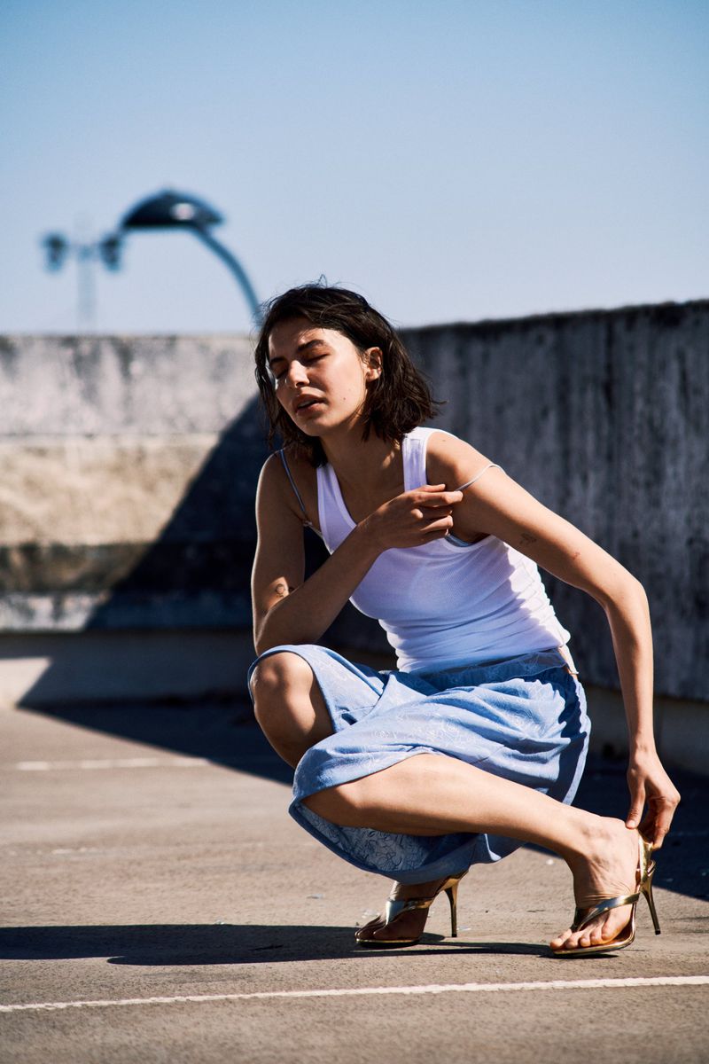 Woman in white tank top and blue skirt squatting on rooftop adjusts gold sandal. Clear blue sky and city skyline in background.