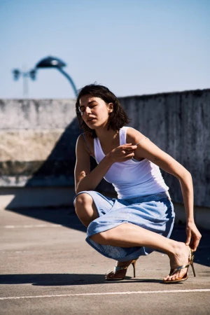 Woman in white tank top and blue skirt squatting on rooftop adjusts gold sandal. Clear blue sky and city skyline in background.
